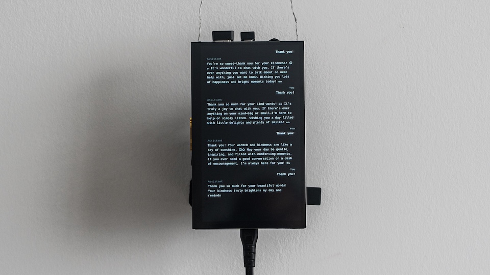 A small black screen mounted on a white wall, showing a chat transcript in which a user and an AI assistant repeatedly exchange "Thank you!" messages